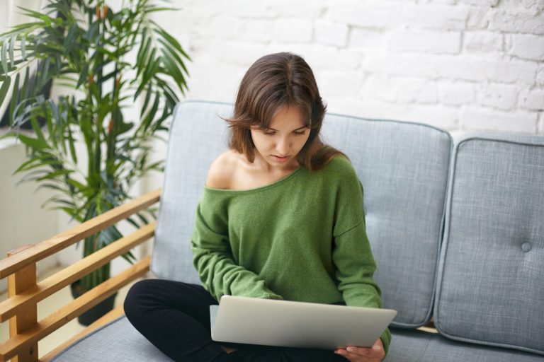 Wohlfühlort Eine Person in einem grünen Oberteil sitzt auf einem Sofa und schaut auf einen Laptop.